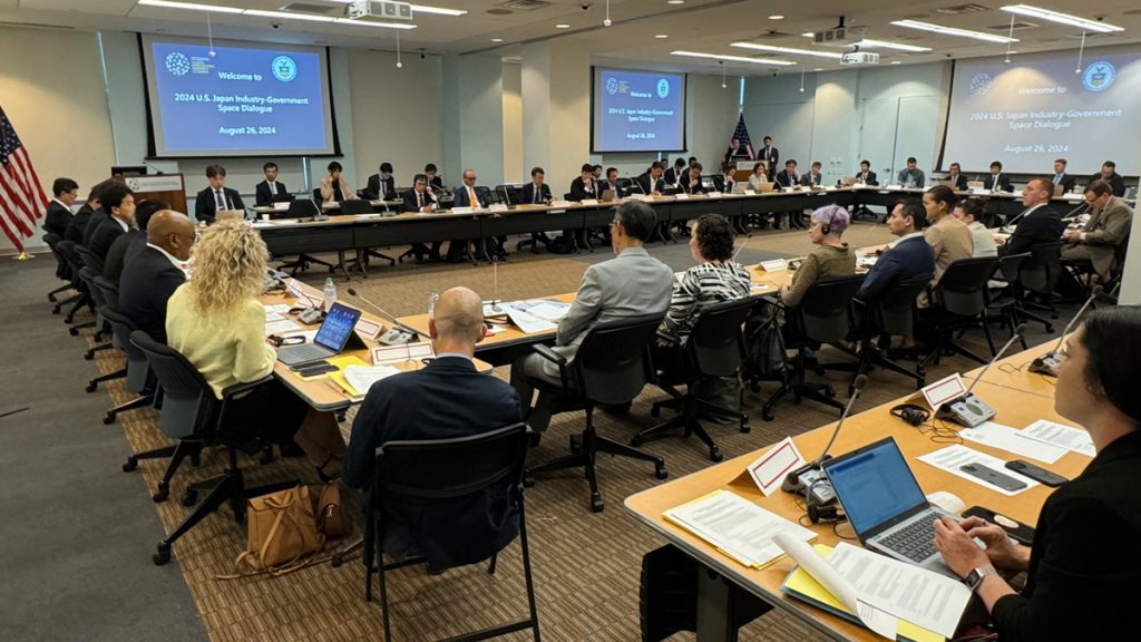 Wide shot of conference room with U.S. and Japanese delegates seated around a long round table. Projection screens on the walls read “Welcome to 2024 U.S. Japan Industry-Government Space Dialogue | August 26, 2024”.