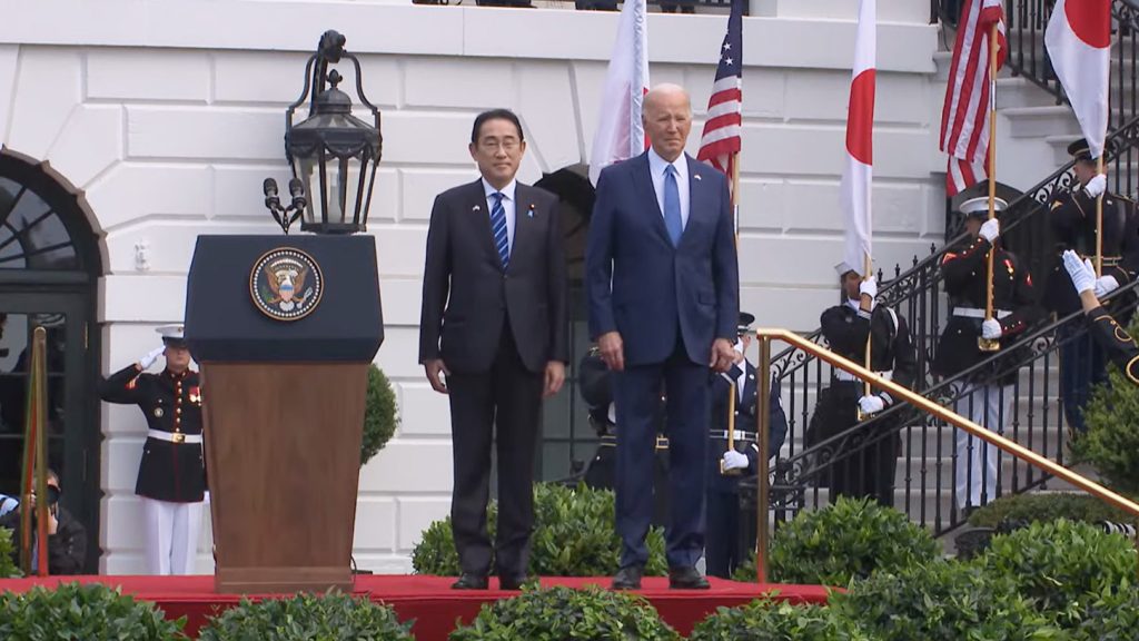 Prime Minister Kishida and President Biden stand together on a podium with uniformed military personnel holding U.S. and Japanese flags behind them