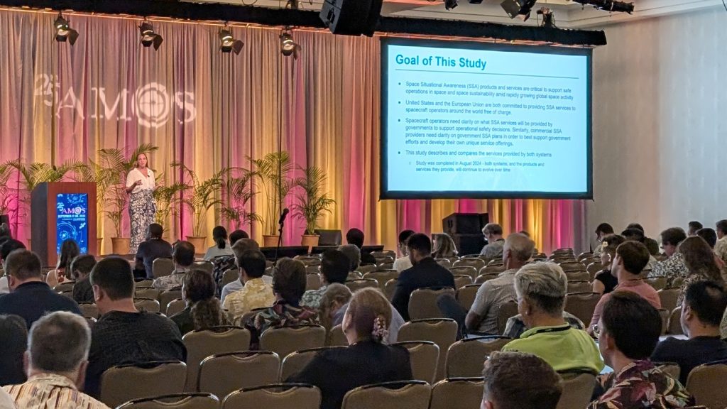 Mariel Borowitz presents on stage with a "25th AMOS" logo, potted palms, and a screen reading, "Goal of This Study." A large audience is seated in front of her.